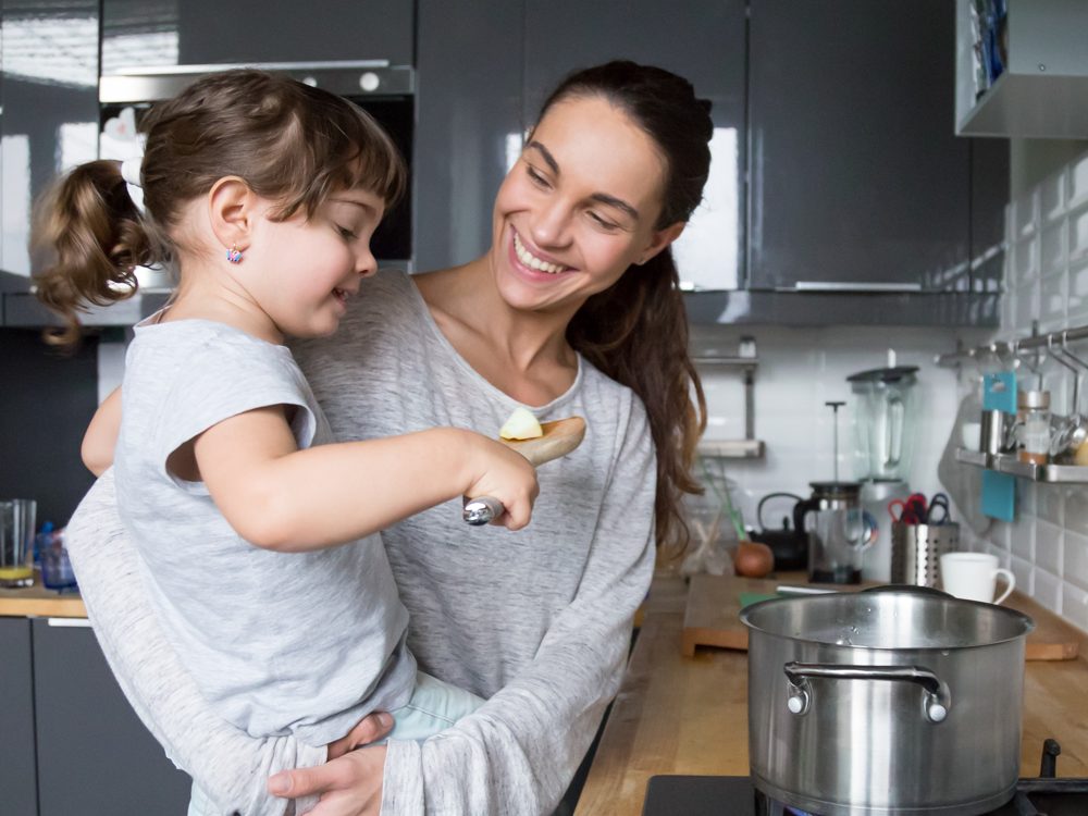 A woman laughing with her young daughter as they cook together at the stove